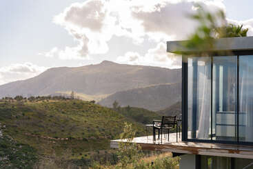 Une maison moderne aux grandes baies vitrées donne sur des collines verdoyantes et vallonnées ainsi que sur les montagnes au loin, sous un ciel parsemé de nuages cotonneux. Un petit salon de jardin invite à la détente.