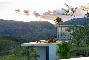 Une maison moderne aux parois de verre se niche sur une colline verdoyante. Deux chaises longues sont disposées sur le toit, d'où l'on peut admirer le panorama montagneux au coucher du soleil, avec des palmiers à proximité.