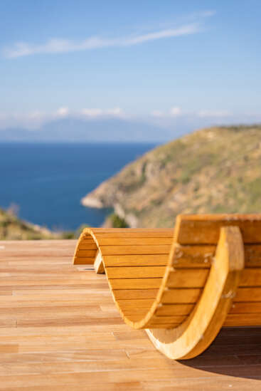 La photo montre un banc en bois élégant aux lignes ondulées, posé sur une terrasse en bois, avec vue sur un paysage côtier pittoresque où se mêlent la mer bleue et les montagnes au loin.