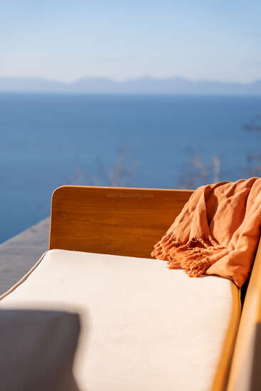 Une chaise longue en bois confortable, agrémentée d'un coussin beige et d'un plaid orange, donne sur une mer bleue paisible et des montagnes lointaines sous un ciel dégagé. Une atmosphère apaisante.