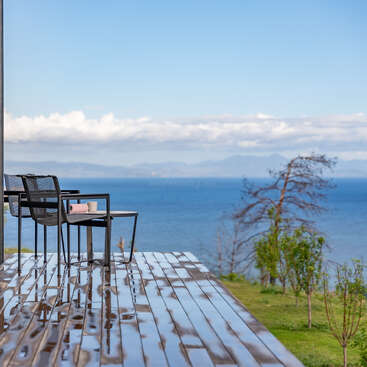 Une terrasse moderne, agrémentée de chaises noires, offre une vue apaisante sur l'océan. Les reflets sur le parquet évoquent la pluie. La végétation et les montagnes au loin viennent parachever ce tableau paisible.