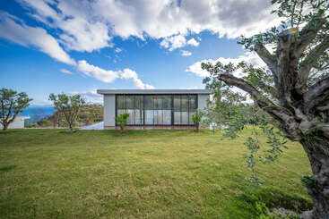 Une maison moderne et minimaliste, dotée de grandes baies vitrées, se dresse sur une pelouse verte, entourée d'arbres, sous un ciel d'un bleu éclatant parsemé de nuages. Un paysage magnifique.
