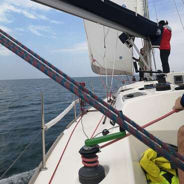 La imagen representa un velero en el agua, con una persona de pie en la cubierta, con una chaqueta roja, y una cuerda que se extiende desde el primer plano hasta el mástil del barco.