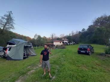 Ein Mann steht lächelnd auf einer Wiese mit Autos, einem Dachzelt, Bäumen und einem Haus im Hintergrund, was eine entspannte Camping-Atmosphäre schafft.
