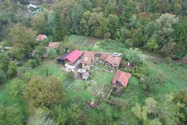 Luftaufnahme eines ländlichen Gehöfts mit mehreren Häusern, roten Dächern, Gartenparzellen, umliegenden Bäumen, grünen Feldern und einem Waldgebiet im Hintergrund. Friedliche Umgebung.