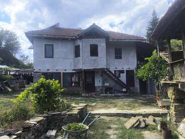 Ein rustikales, zweistöckiges Haus mit einem roten Ziegeldach und einer Außentreppe steht in einem ländlichen Hof, umgeben von Grün, Steinmauern und Gartengeräten.