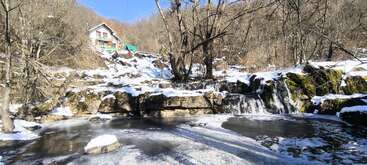 Eine ruhige Winterszene zeigt einen teilweise gefrorenen Wasserfall, einen ruhigen Bach, blattlose Bäume und ein Haus auf einem schneebedeckten Hügel unter klarem, blauem Himmel.