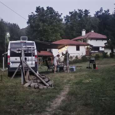 Ein Wohnmobil ist in der Nähe eines Hauses geparkt. Zwei Leute bauen unter einer Markise die Außenbeleuchtung auf und bereiten sich auf ein abendliches Treffen auf einer Wiese vor.
