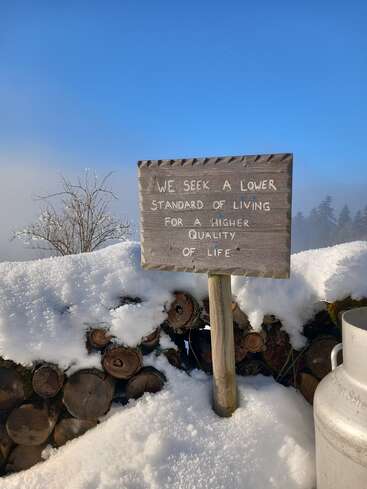 A wooden sign in snowy outdoors reads: “We seek a lower standard of living for a higher quality of life.” Logs and a milk can nearby, clear sky above.