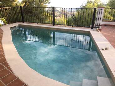 Ein kleiner, sauberer, rechteckiger Swimmingpool mit Stufen, umgeben von einer gefliesten Terrasse und einem schwarzen Metallzaun. Hinter dem Zaun sind Bäume und entfernte Hügel zu sehen.