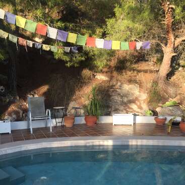 Una apacible escena junto a la piscina con coloridas banderas de oración, plantas en macetas, una tumbona y árboles de fondo crean un entorno exterior tranquilo y lleno de naturaleza.