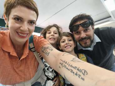 A family of four smiles for a selfie. The woman in front has tattoos and wears orange, with two children and a man with sunglasses in the background.