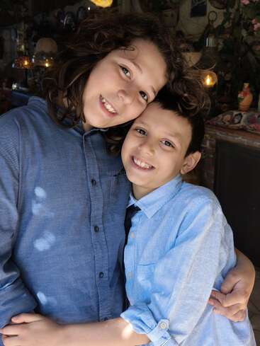 Deux enfants souriants, vêtus de chemises bleues, se tiennent serrés l'un contre l'autre, les bras autour des épaules, rayonnant d'affection et de chaleur dans un intérieur chaleureux et bien éclairé.