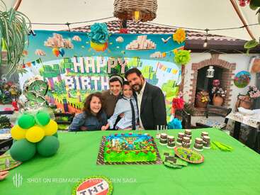 A family celebrates a child’s Minecraft-themed birthday party. Colorful decorations, a cake, balloons, and happy faces fill the festive scene under a decorated canopy.