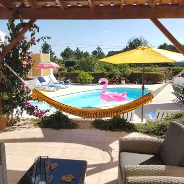 The image depicts a serene backyard oasis, featuring a pool, hammock, and patio furniture, surrounded by lush greenery and vibrant flowers on a sunny day.