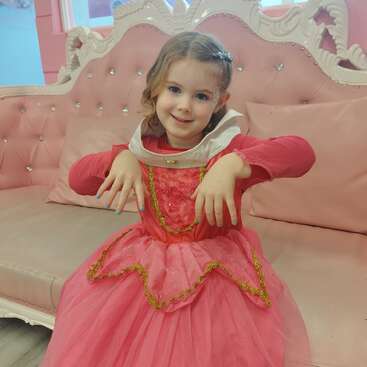 A imagem mostra uma menina com um vestido de princesa rosa, sentada em um sofá rosa, com as mãos apoiadas nos joelhos, exibindo as unhas pintadas.