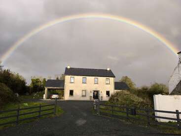 Une maison à deux étages se dresse sous un ciel dramatique et nuageux, magnifiquement encadré par un arc-en-ciel vibrant au-dessus de la tête. Une verdure luxuriante et une voiture complètent la scène rurale.