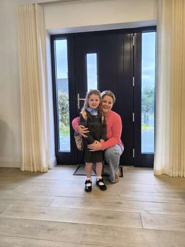 Une femme est agenouillée à côté d'une jeune fille en uniforme scolaire, souriante devant une porte d'entrée noire, prête pour la journée d'école, en intérieur avec un éclairage lumineux.