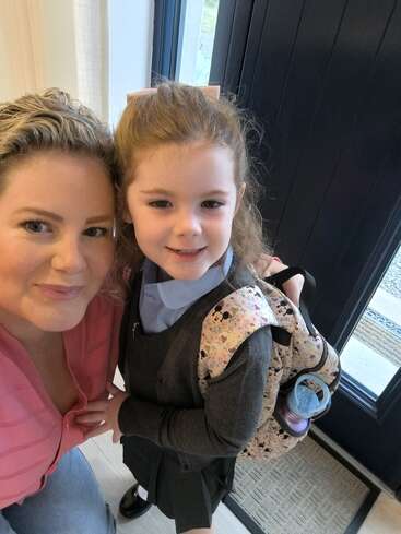 Une femme et une jeune fille sourient ensemble à l'intérieur. La jeune fille porte un uniforme d'écolière, un sac à dos et un nœud, ce qui laisse supposer qu'il s'agit de son premier jour d'école.