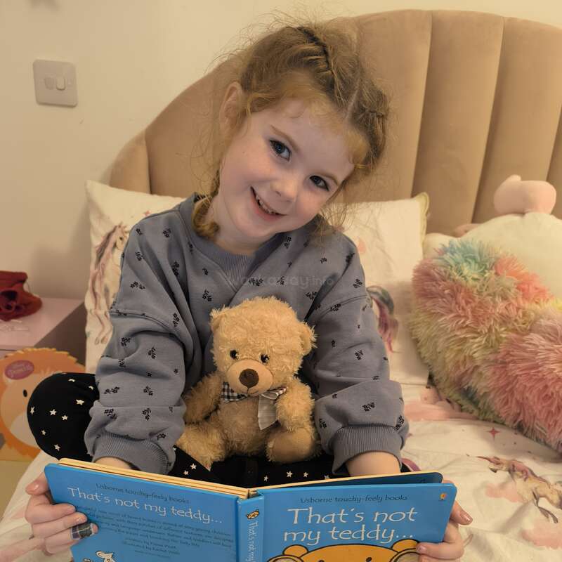 Une jeune fille est assise sur son lit, souriante, tenant un ours en peluche et lisant "That's not my teddy" (ce n'est pas mon ours en peluche). Des oreillers douillets et colorés complètent cette scène chaleureuse.