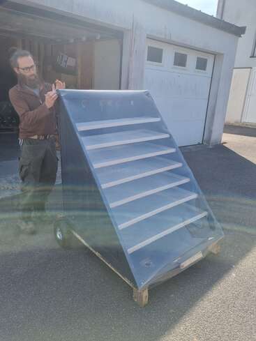 A bearded man stands near a homemade solar dehydrator with transparent cover and wooden shelves, positioned outdoors on wheels in front of a garage. Sunlight streaks across.