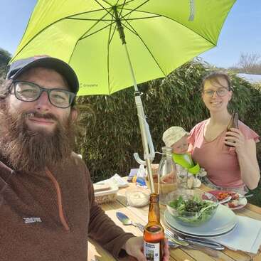Un hombre con barba, una mujer y un bebé disfrutan de una comida al aire libre bajo una sombrilla verde. Sonríen, con bebidas en la mano, rodeados de comida, ensalada y rayos de sol. Ambiente relajado.