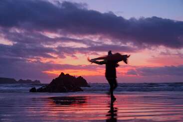 Un surfeur marche le long d'une plage aux reflets chatoyants au coucher du soleil, sa planche de surf à la main. Des nuages spectaculaires, des formations rocheuses et des couleurs vives créent une atmosphère à la fois sereine et pleine d'aventure.