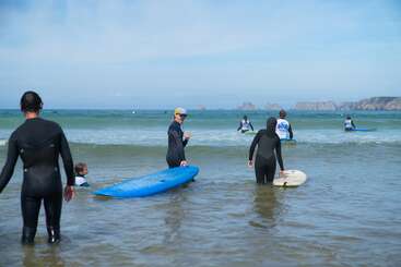 Plusieurs personnes en combinaison de plongée se trouvent sur une plage, une planche de surf à la main, debout dans l'eau peu profonde, s'apprêtant à surfer. On aperçoit l'océan et des falaises au loin.