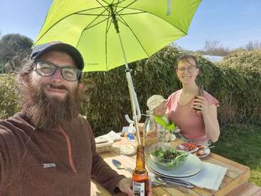 A bearded man, woman, and baby enjoy an outdoor meal under a green umbrella. They smile, holding drinks, surrounded by food, salad, and sunshine. Relaxing atmosphere.