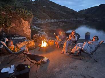 Quatro pessoas sentadas ao redor de uma fogueira acesa à beira de um lago calmo, assando marshmallows, enroladas em cobertores, desfrutando de uma noite aconchegante ao ar livre cercada pela natureza.