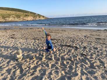 Uma criança alegre em uma roupa de mergulho azul posa energicamente em uma praia de areia ao lado de pequenas torres de pedra empilhadas, com ondas e colinas gramadas ao fundo.