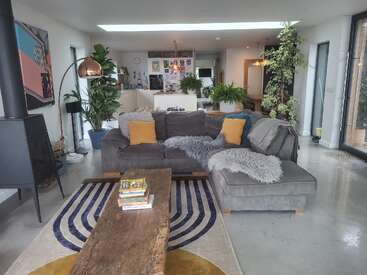 Sala de estar moderna com sofá seccional cinza, almofadas coloridas, mesa de centro de madeira, plantas de interior, cozinha aberta ao fundo, decoração aconchegante e iluminação natural.