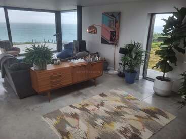 Uma sala de estar moderna com grandes janelas oferece vistas deslumbrantes do oceano. Móveis elegantes, plantas exuberantes, um tapete estampado e decoração contemporânea criam uma atmosfera aconchegante e convidativa.