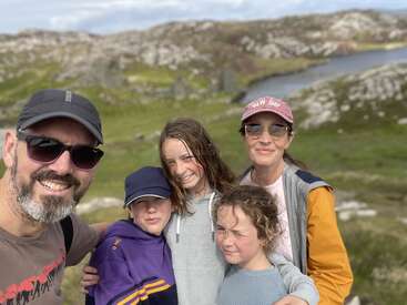 Uma família sorridente de cinco pessoas posa ao ar livre em uma paisagem rochosa cênica com colinas verdes e um rio. Eles parecem felizes, usando roupas casuais e chapéus.