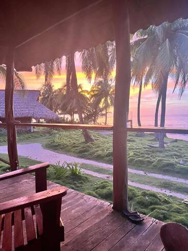 The image shows a peaceful tropical scene with palm trees, a wooden porch, chairs, a hammock, and a beautiful pastel sunset over the ocean. Relaxing atmosphere.