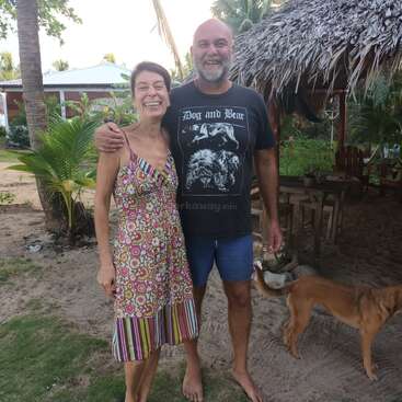 A smiling man and woman stand barefoot on sandy ground outdoors, with a dog nearby, tropical plants, thatched hut, and relaxed, tropical atmosphere in the background.