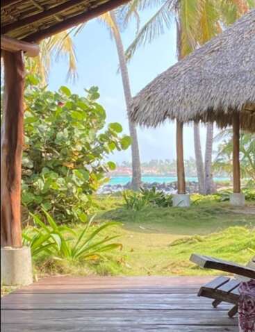 A tropical scene with palm trees, wooden patio, thatched-roof hut, and vibrant greenery overlooks the blue ocean, creating a peaceful and relaxing beach atmosphere.