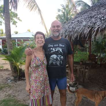 Un homme et une femme souriants se tiennent pieds nus sur un sol sablonneux en plein air, avec un chien à proximité, des plantes tropicales, une hutte au toit de chaume et une atmosphère tropicale détendue en arrière-plan.