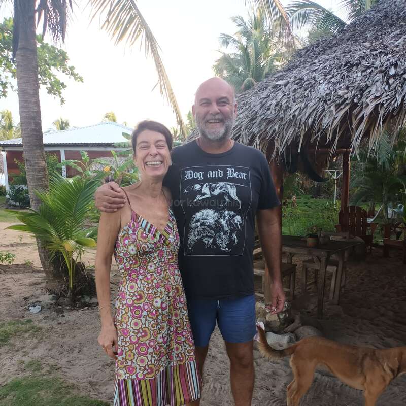 A smiling man and woman stand barefoot on sandy ground outdoors, with a dog nearby, tropical plants, thatched hut, and relaxed, tropical atmosphere in the background.