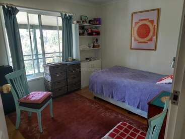 This cozy bedroom features a single bed with a purple cover, shelves, wicker drawers, two chairs, a large window with curtains, and a bright red rug.