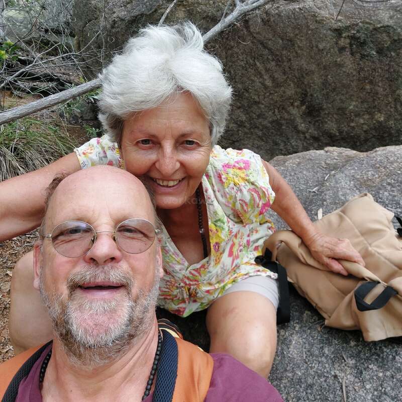 A smiling older couple takes a selfie outdoors while hiking. They are sitting on rocks, surrounded by nature, appearing happy and relaxed on their adventure together.