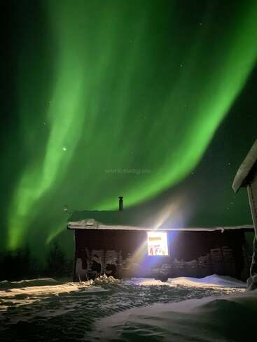 Brilliant green northern lights dance across a dark winter sky above a snow-covered cabin, warmly lit from within, creating a serene and magical Arctic scene.
