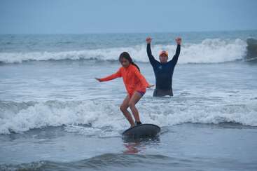 Uma menina com uma camisa laranja se equilibra em uma prancha de surf nas ondas rasas do mar, enquanto um homem atrás dela torce com os braços levantados em sinal de incentivo.