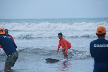 Um jovem surfista com uma camisa laranja treina ficar em pé na prancha nas ondas rasas do mar, cercado por treinadores vestindo camisas azuis e chapéus laranja.