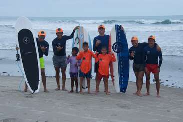 Um grupo de crianças e adultos posa na praia com pranchas de surf, usando chapéus e camisetas combinando, sorrindo e gesticulando, com as ondas do mar ao fundo.