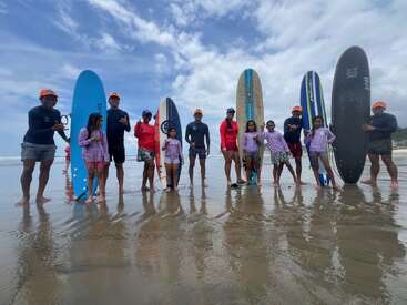 Um grupo de adultos e crianças está em uma praia, cada um com uma prancha de surf nas mãos. Eles estão usando camisetas de proteção solar e chapéus, sorrindo sob um céu azul nublado.