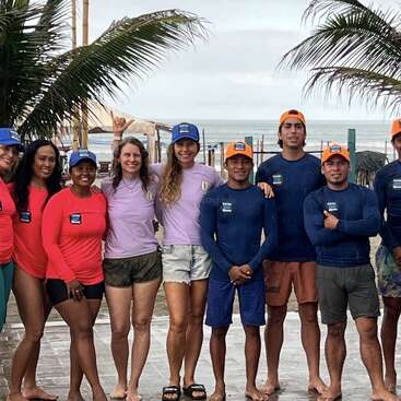 A group of people poses together outdoors near a beach, wearing colorful shirts and hats. Palm trees frame them, and the ocean is visible in the background.