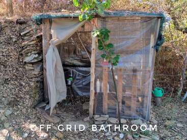 Esta imagen muestra un baño improvisado al aire libre construido con madera, láminas de plástico y palés. Rodeado de naturaleza, en la parte inferior pone "OFF GRID BATHROOM".