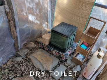 Esta imagen muestra un retrete seco improvisado en un refugio rústico al aire libre, con un asiento de plástico, artículos para lavarse las manos, estanterías, piedras y un suelo cubierto de hojas.