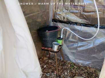 Esta imagen muestra una ducha improvisada al aire libre: una tienda de plástico, un cubo negro, una taza de metal, un taburete, una bomba eléctrica y un texto recordatorio para calentar primero el agua.
